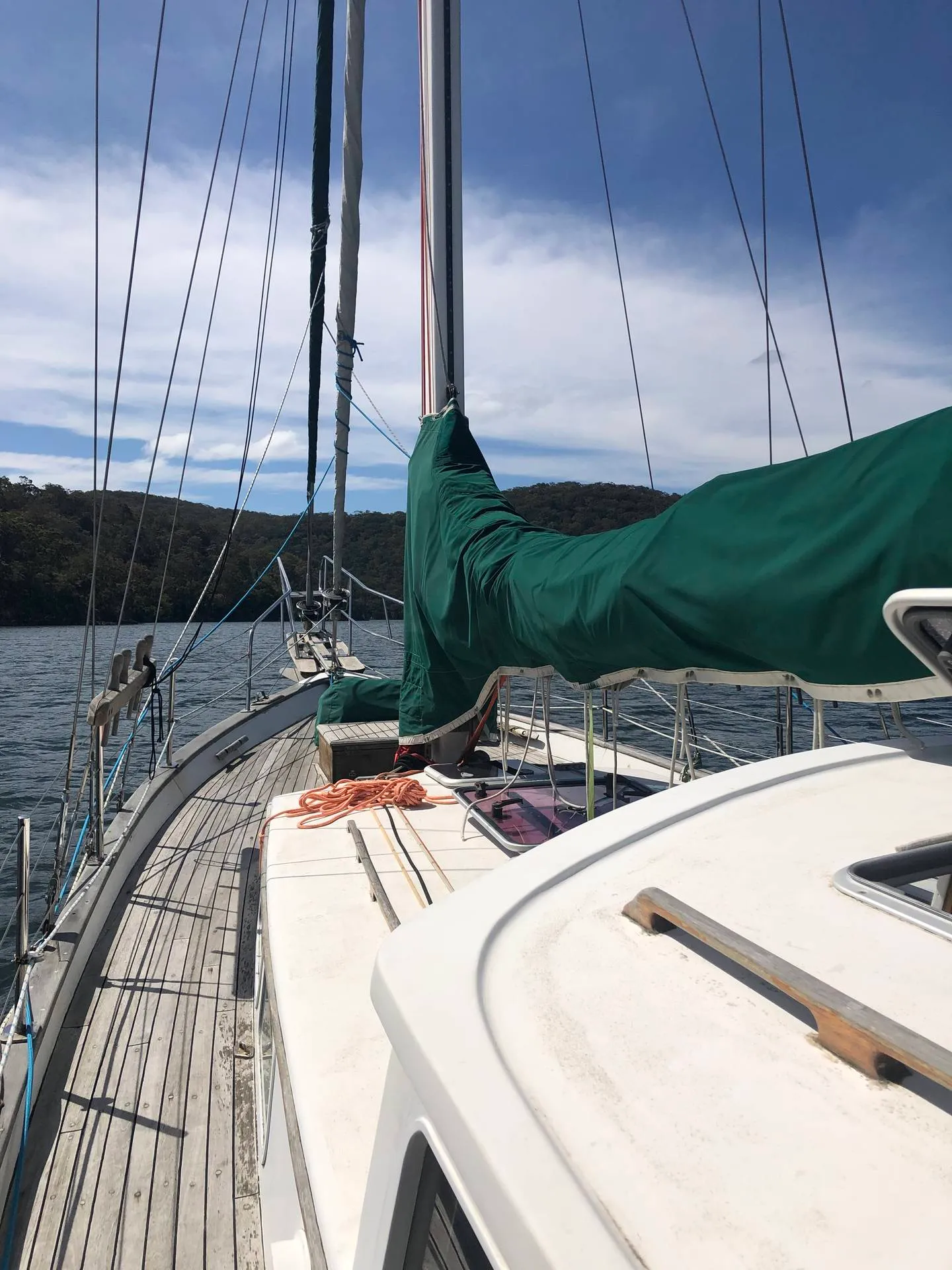 Looking forward along the deck of the sailing boat we live aboard — sail cover on the boom, timber side decks, and bush-covered river shoreline beyond.