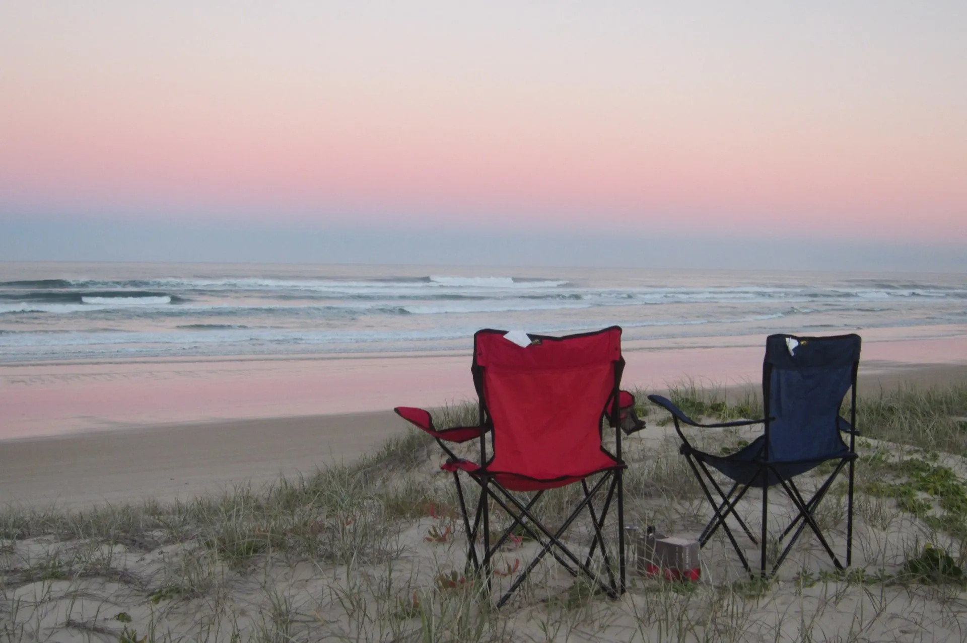 Two camp chairs side by side on a dune above a remote surf beach at dusk.